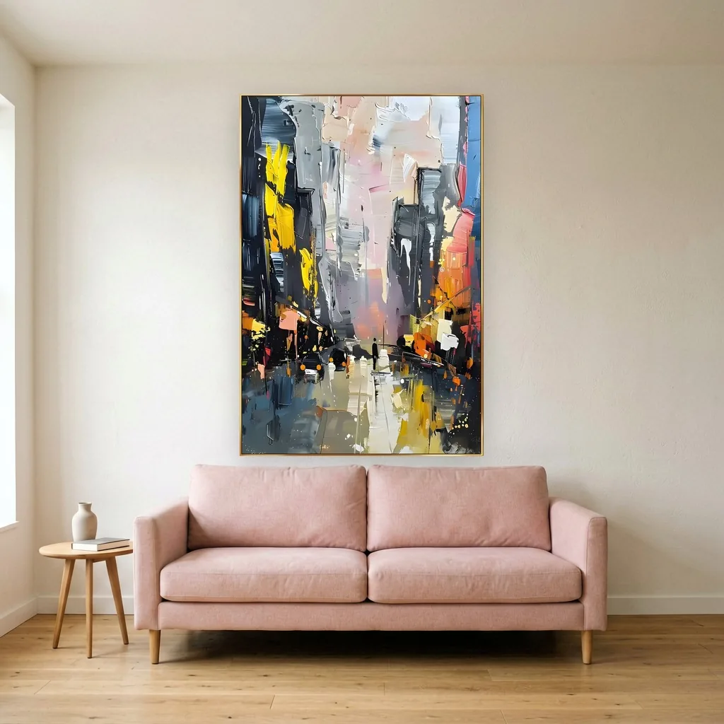 Rainy dusk street where a lone figure walks between charcoal buildings lit with yellow windows, a palette-knife oil city canvas on a cream wall above a blush pink sofa and a small round side table