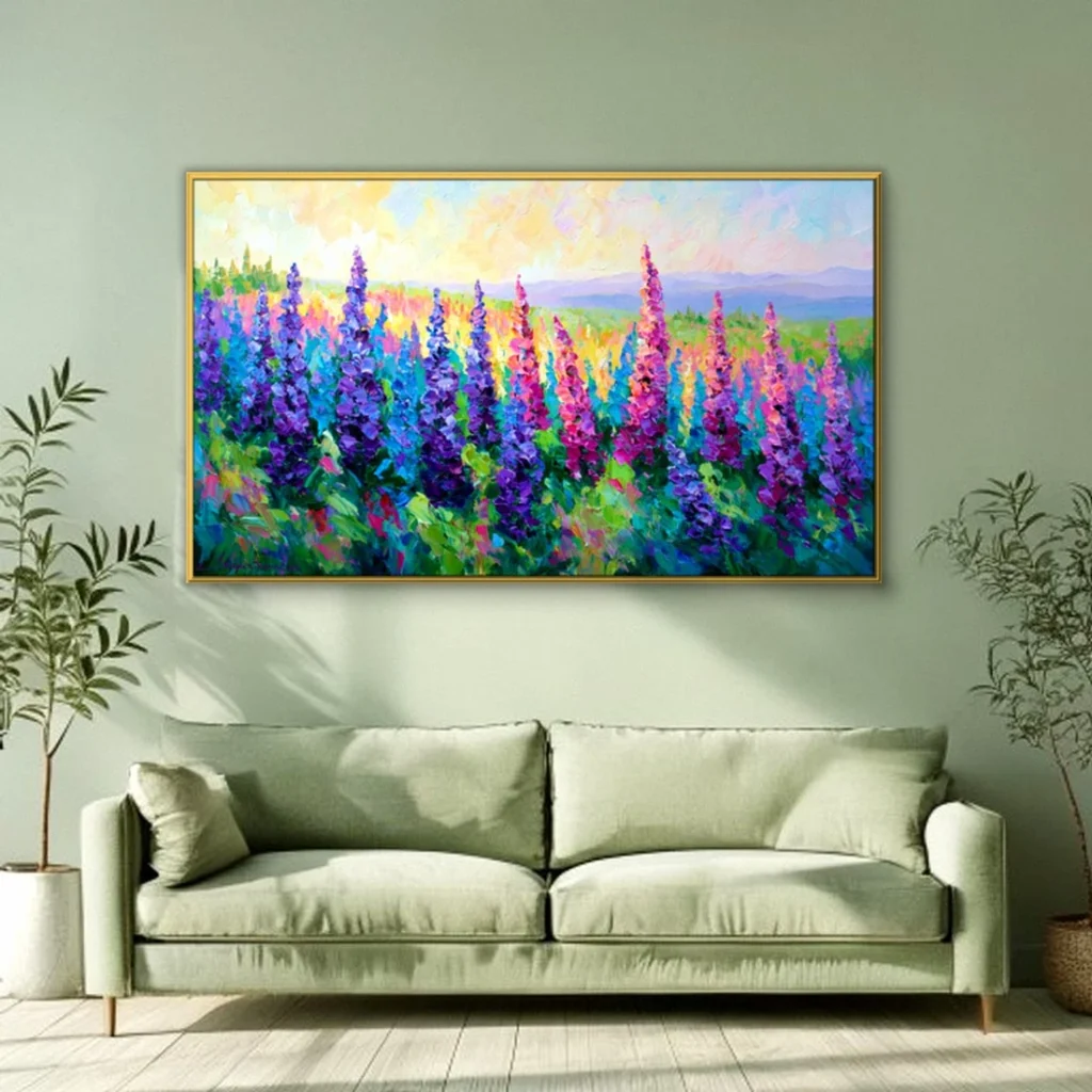 Wide palette-knife oil painting of purple and pink flower spires above a bright meadow under a warm sunset sky, framed in gold on a sage-green wall above a light green linen sofa between two potted trees.