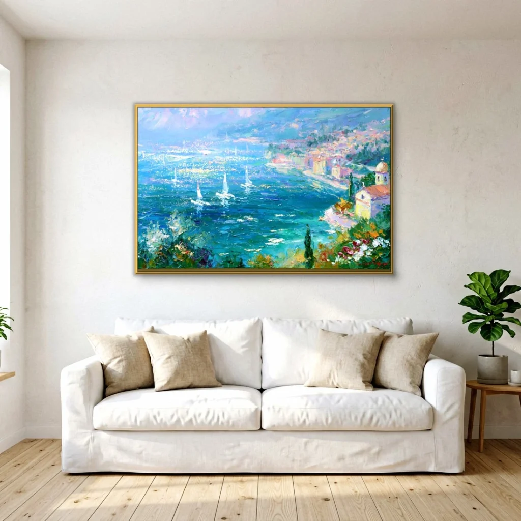 Gold-framed Mediterranean seascape painting of a blue bay with white sailboats and a pale coastal village, hung on a white wall above a cream slipcovered sofa with linen pillows and a potted plant on light plank floor.